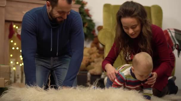 Mother, father and little baby sitting on the floor in the room with christmas decoration. Man gives small present box to child crawling on fluffy carpet. Happy family celebrating Christmas together - Filmagem, Vídeo