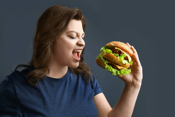 Plus size woman with tasty burger on grey background. Concept of body positive - Фото, изображение