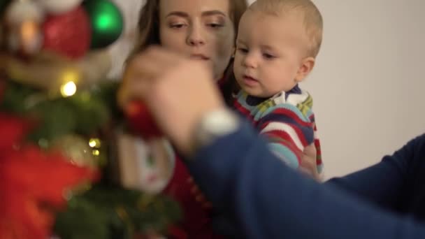 Mother, father and little baby decorating new year tree close-up. Woman holding child near fir tree, showing bright decoration, man hanging toys. Happy family celebrating Christmas together - Video