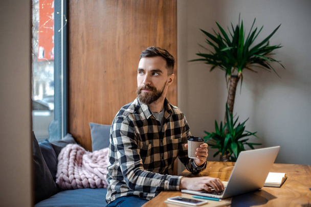 Felnőtt stílusos szakállas csípő ember ül az ablak mellett az utcai város Cafe Shoo, dolgozó online Internet marketing a számítógépén, elfoglalt és koncentrált koncepció - Fotó, kép