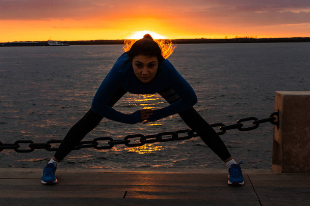 Gün batımı ve Amur nehrine karşı doğada spor egzersizleri yapan spor kıyafetler içinde çekici bir kadın, jimnastik seviyor, bacaklarını yoğurur. Aktif genç kız spor yapan, sağlıklı yol açar - Fotoğraf, Görsel
