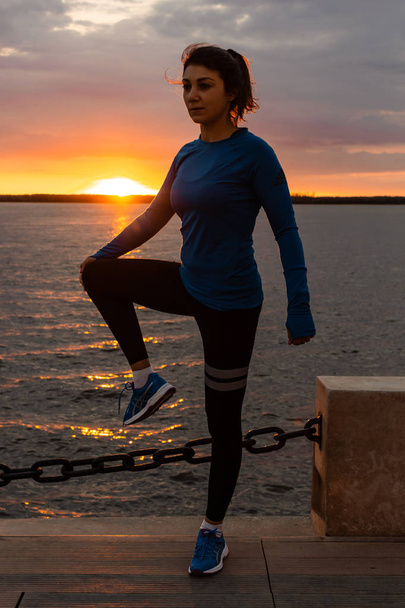 Khabarovsk, Rusia - 07 / 10 / 2018: Una mujer atractiva en ropa deportiva haciendo ejercicios deportivos en la naturaleza contra el atardecer y el río Amur, ama la gimnasia, amasa sus piernas. Chica joven activa
 - Foto, imagen
