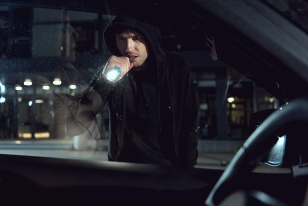 thief in black hoodie looking into car with flashlight - Zdjęcie, obraz