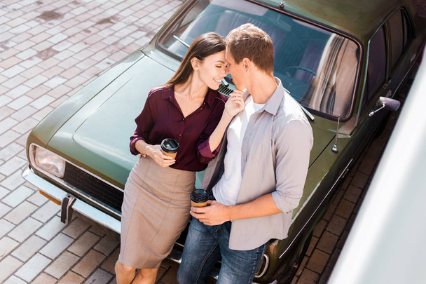 Beautiful carefree couple discovering the world traveling by car. Young man and woman kissing, hugging and touching each other while sitting in hood of the car, green retro car background. - Foto, immagini