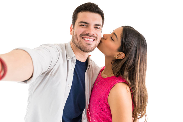 Beautiful woman kissing cheerful handsome boyfriend while taking selfie with him against plain background - Photo, Image