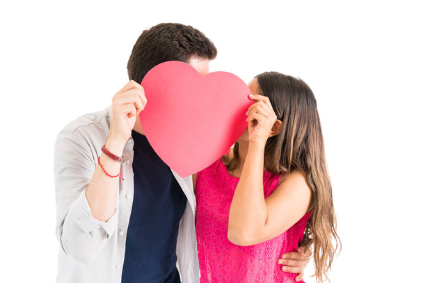 Jovem casal apaixonado beijando enquanto esconde o rosto atrás de um grande coração de papel vermelho contra fundo branco
 - Foto, Imagem