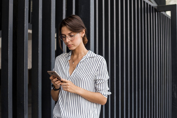 Сконцентрированная женщина отправляет сообщения с использованием современных приложений на мобильный телефон во время прогулки по городской улице, свободное время для общения по современным технологиям на открытом воздухе, используя 5G Интернет
 - Фото, изображение