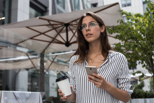 Pozitivní módní student s mobilním telefonem v ruce a občerství kávu, která se nachází nedaleko kavárny, šťastná žena se usmívá a dívá se na čekající aktualizační oznámení na mobil pomocí Wree Fiwi čekající na taxi - Fotografie, Obrázek