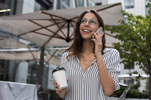 Gorgeous female lecturer of university calls via cell telephone and holds coffee standing by street cafe, coffee break, young woman office worker is having mobile phone conversation during work break - Photo, Image