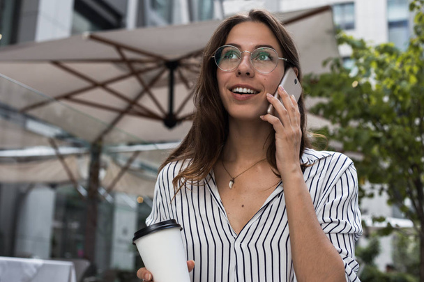 Happy woman enjoying lifestyle outdoors during coffee break and positive smartphone conversation, cheerful hipster girl calling to customer service feeling excited of information about account balance - Фото, изображение