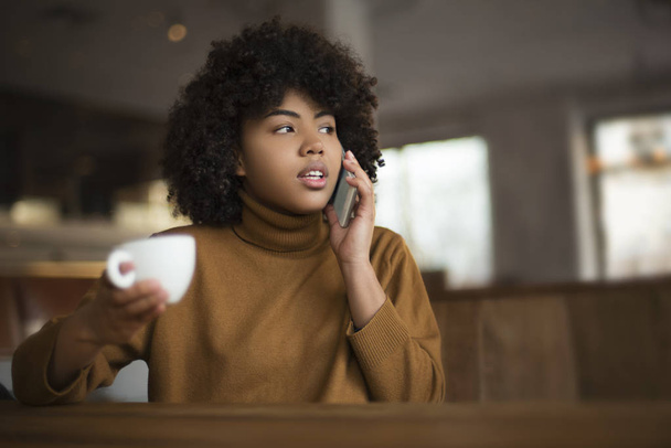 A young girl of African-American appearance sits at a table. She has a coffee break in a warm uninhabited atmosphere. She talks on the phone with her friends, has a rest and drinks tasty coffee - Photo, image