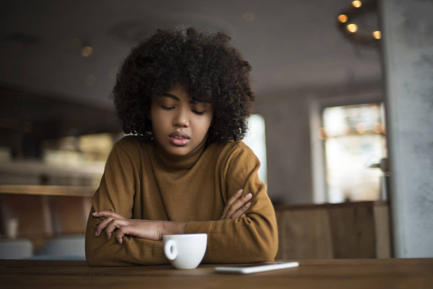 Ένα νεαρό κορίτσι με Αφρο-Αμερικανική εμφάνιση κάθεται σε ένα τραπέζι. Έχει διάλειμμα για καφέ σε μια ζεστή ακατοίκητη ατμόσφαιρα. Μιλάει στο τηλέφωνο με τους φίλους της, έχει ξεκούραση και πίνει νόστιμο καφέ. - Φωτογραφία, εικόνα