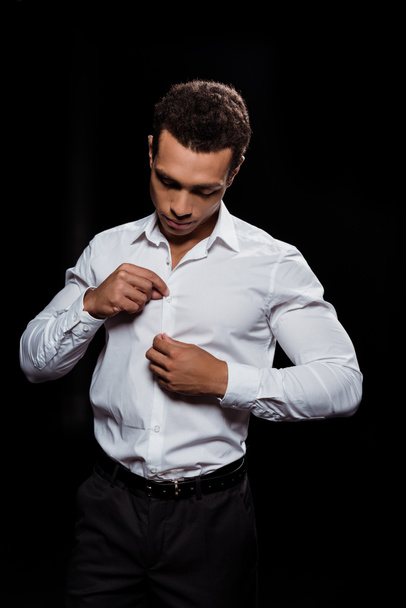handsome bi-racial man touching and looking at white shirt isolated on black  - Foto, Bild