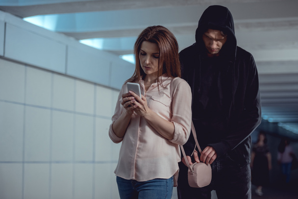 ladrón robar dinero de la bolsa de la mujer usando el teléfono inteligente en el paso subterráneo
 - Foto, imagen