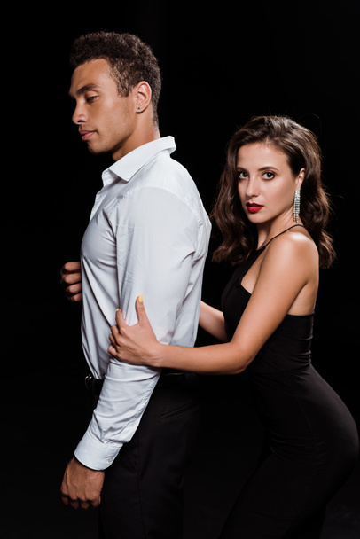 elegant woman hugging handsome mixed race man looking at camera isolated on black  - Photo, Image