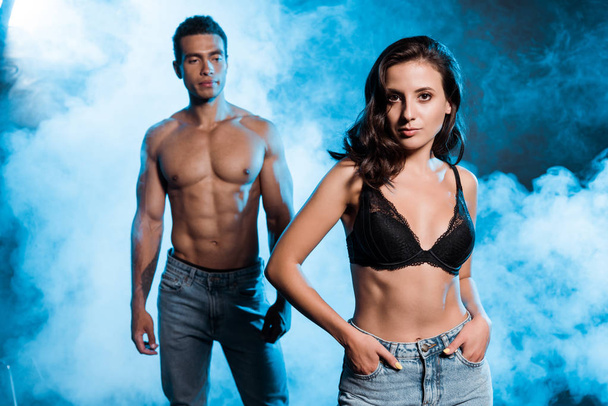 selective focus of attractive girl standing with hands in pockets near handsome bi-racial man on blue with smoke  - 写真・画像