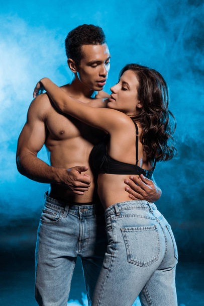 sexy mixed race man hugging and looking at girl in bra and jeans on blue with smoke  - Fotó, kép