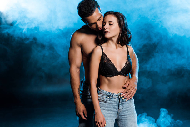 handsome mixed race man hugging attractive girl in lace black bra on black with blue smoke  - Фото, зображення