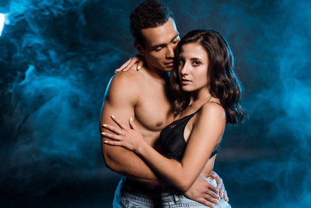 muscular bi-racial man hugging young woman in jeans and bra on black with smoke  - Photo, Image