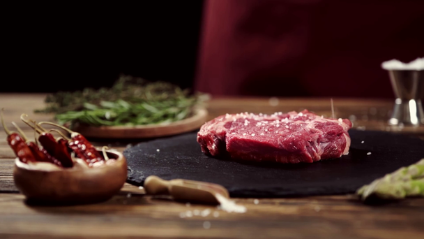 Cropped view of man seasoning meat steak with salt near ingredients on table isolated on black - Metraje, vídeo