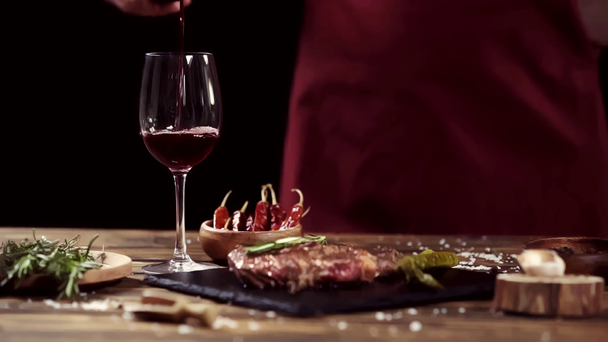 geschnittene Ansicht eines Mannes, der Wein in Glas gießt, in der Nähe von Fleischsteak und Zutaten auf einem Tisch isoliert auf schwarz - Filmmaterial, Video