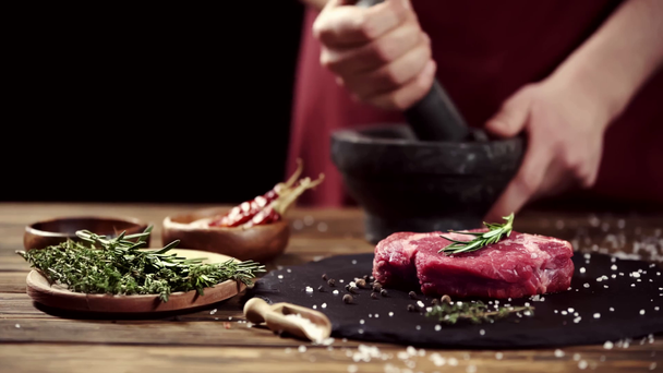 Vista ritagliata dell'uomo macinare spezie con pestello e malta vicino carne cruda bistecca e ingredienti sulla tavola isolata su nero
 - Filmati, video
