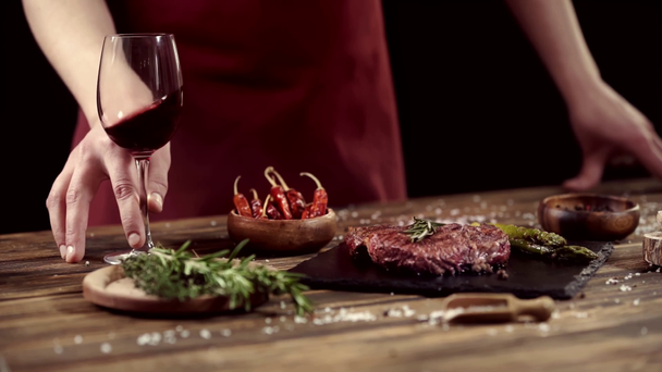 Oříznutý pohled člověka na míchání skleněného vína v blízkosti masových steaků a přísad na stůl izolovaný na černém - Záběry, video