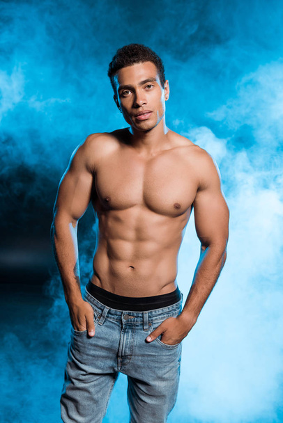 sexy mixed race man standing with hands in pockets and looking at camera on blue with smoke  - Foto, afbeelding