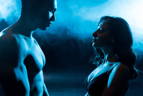 vue latérale de la jeune femme en soutien-gorge regardant sexy musclé mixte race homme sur bleu avec de la fumée
  - Photo, image