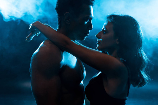 side view of young attractive woman in bra hugging sexy muscular mixed race man on blue with smoke  - Photo, image