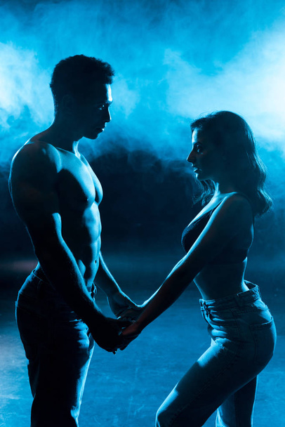 side view of young woman in bra holding hands with sexy muscular man on blue with smoke  - 写真・画像