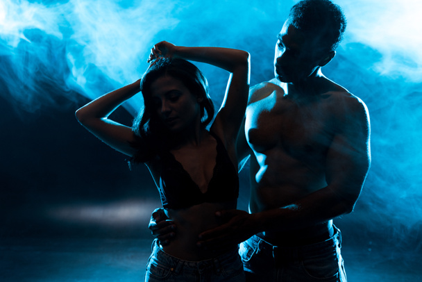 sexy shirtless mixed race man looking at attractive girl on blue with smoke  - 写真・画像