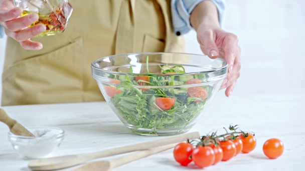 テーブルの上の食材の近くに野菜サラダにオリーブオイルを注ぐ女性のトリミングビュー - 映像、動画
