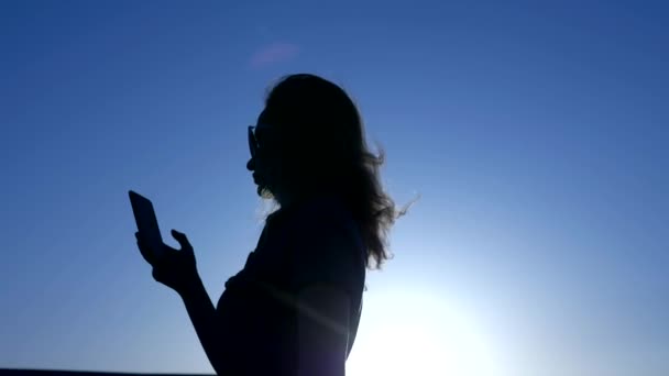 Silueta ženy s smartphone s letíma vlasama na pozadí modrého nebe slunce - Záběry, video