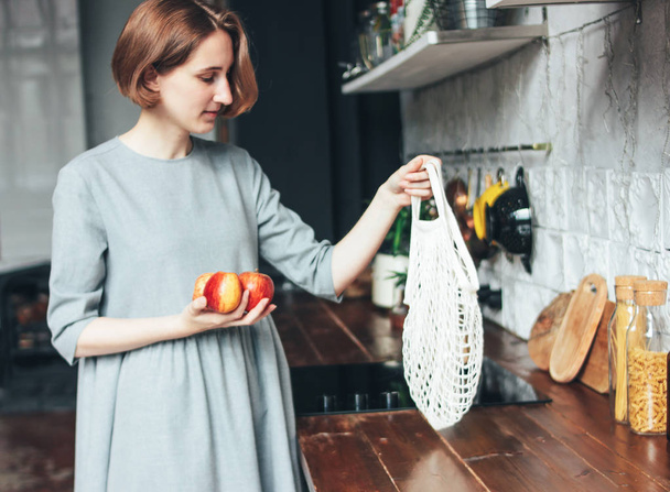 Jovem mulher em vestido cinza puxa maçãs de malha saco de pano saco de corda comprador na cozinha, zero desperdício, vida lenta
 - Foto, Imagem