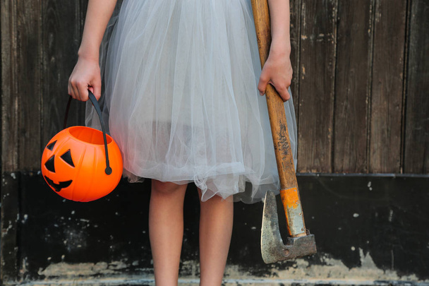 Glimlachend blond meisje houdt Halloween pompoen Jack O'Lantern en een bijl in handen op houten achtergrond. Halloween viering, horror concept. - Foto, afbeelding