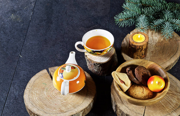 On a dark wooden background are round saw cuts of a tree, a mug with tea and a kettle, a candle, wooden cups, cookies, an apple - Foto, Imagem