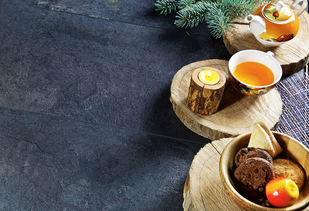 On a dark wooden background are round saw cuts of a tree, a mug with tea and a kettle, a candle, wooden cups, cookies, an apple - Фото, зображення