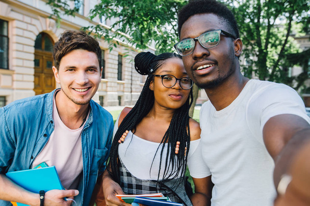 Educazione, stile di vita, tempo libero e tecnologia concettuale.Bello studente africano in occhiali con due amici multietnici che fanno selfie insieme nel parco durante il relax tempo libero
. - Foto, immagini