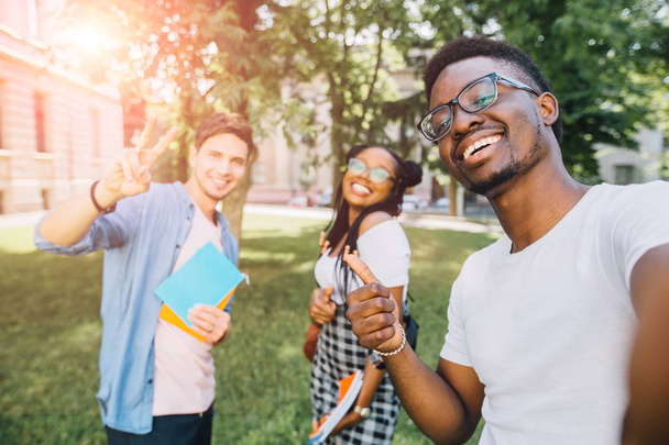 Education,leisure and technology concept. Cheerful friends from different countries and races taking selfie with back lighting. Happy youth people having fun in university campus. Sun glare effect. - Photo, image