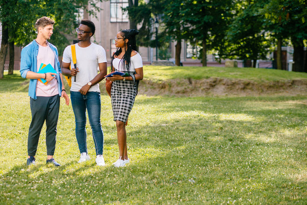 in voller Länge fröhliche multiethnische Studenten, die sich in der Pause an einem sonnigen Sommertag auf dem Campus im Freien unterhalten. Kopierraum. Unterschiedliches Kulturfreundschaftskonzept - Foto, Bild