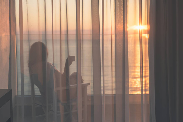 Naked beautiful female wrapped in a blanket sitting on a chair with cup of coffee behind the transparent curtains on the glass balcony early morning at sunrise. Sea view - Fotografie, Obrázek