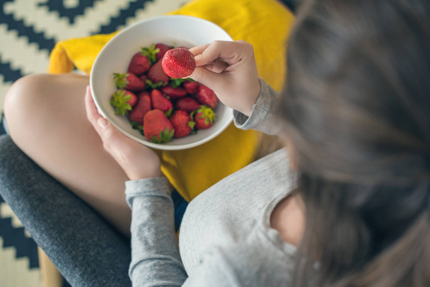 Top view on the woman sitting in the home armchair with ripe strawberry in a bowl. - Fotoğraf, Görsel