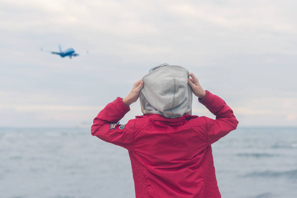 A boy in a red jacket  looks at the sea waves in stormy rainy weather. Landing plane above sea waves in stormy rainy weather. - Фото, зображення