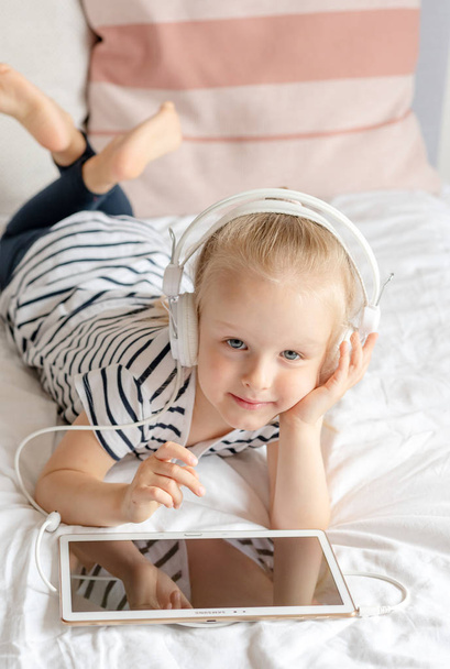 Valkoihoinen pieni tyttö kuulokkeiden katselussa Tablet sängyssä, sisustus, moderni laite Technologies skandinaavinen tyyli
 - Valokuva, kuva
