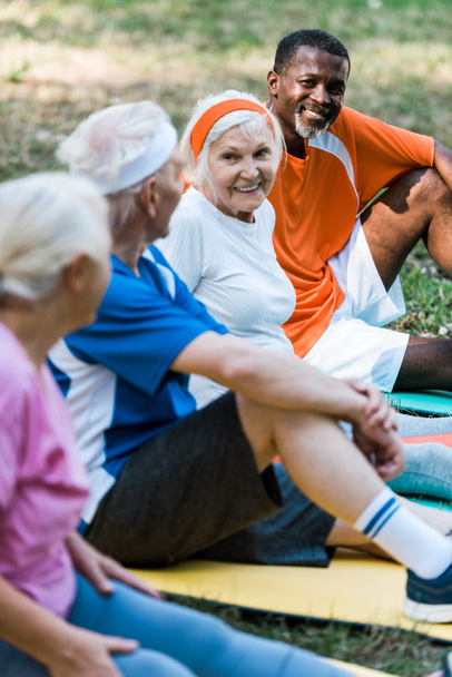 enfoque selectivo de hombres y mujeres multiculturales alegres en ropa deportiva sentado en alfombras de fitness
  - Foto, Imagen