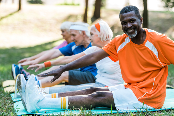 enfoque selectivo de hombre afroamericano feliz cerca de jubilados pensionistas en ropa deportiva ejerciendo en colchonetas de fitness
  - Foto, Imagen
