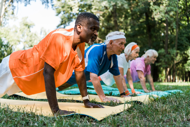 enfoque selectivo del hombre afroamericano haciendo ejercicio de tablón con pensionistas jubilados en colchonetas de fitness
  - Foto, imagen