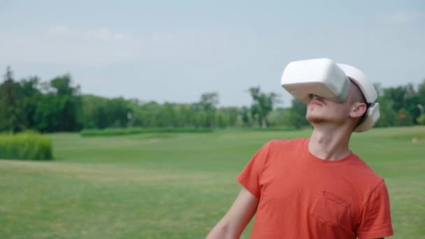 Een man die een vr-headset in het park gebruikt, obstakels ontwijkt en rondkijkt. Een jonge kerel in een rood T-shirt en een helm ervaart Virtual Reality tegen de achtergrond van de natuur. Een mens aan de rechterkant in het frame. 4k-beeldmateriaal. - Video