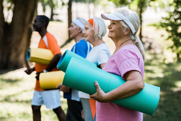 selektivní zaměření šťastné ženy v důchodu v držení fitness rohože poblíž multikulturních důchodců v sportovní přísaze  - Fotografie, Obrázek